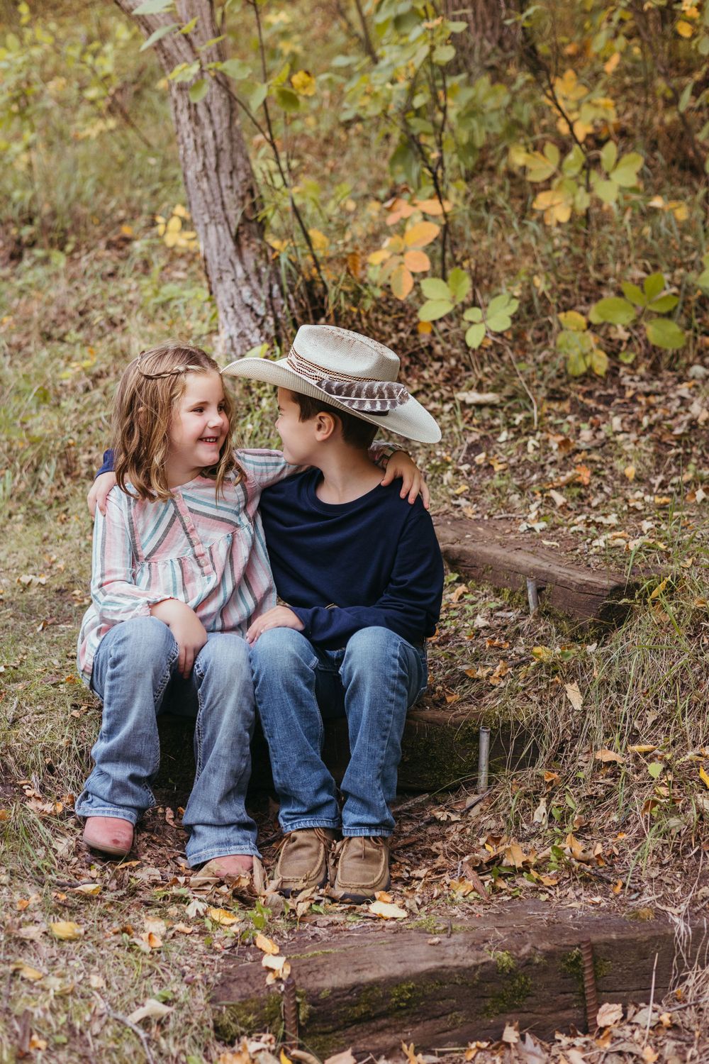 Two people sitting on wooden steps surrounded by autumn foliage share a sweet moment in casual outdoor wear.
