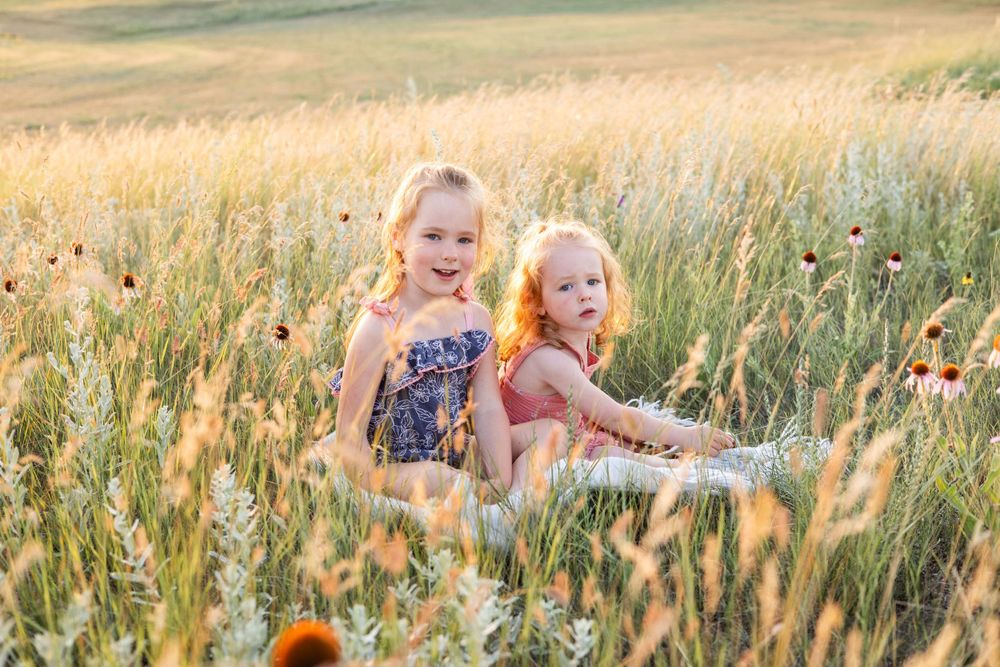 Two children sit together in a field of tall grass and wildflowers during golden hour.