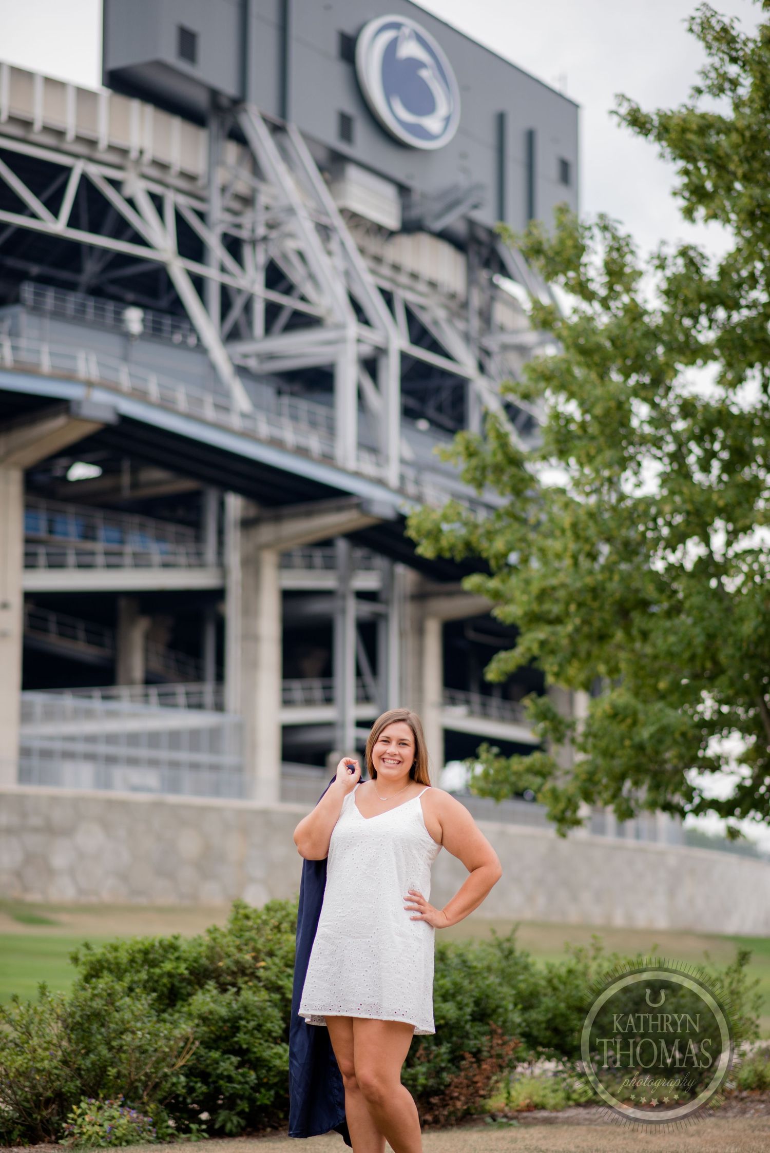 A graduate poses in front of Penn State's Beaver Stadium with cap and gown draped over arm.