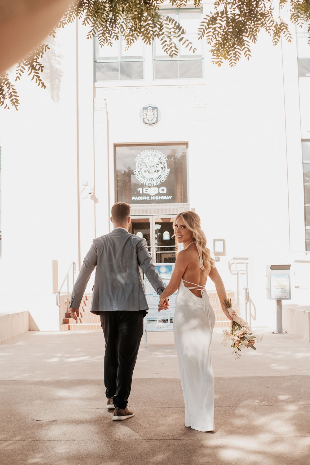 Wedding couple walking hand in hand towards entrance of white building with overhead foliage.