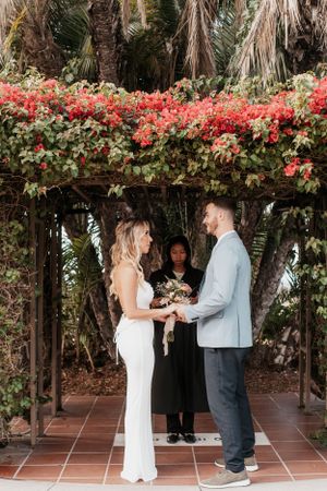 Wedding ceremony under a floral arch with red bougainvillea flowers and palm trees.