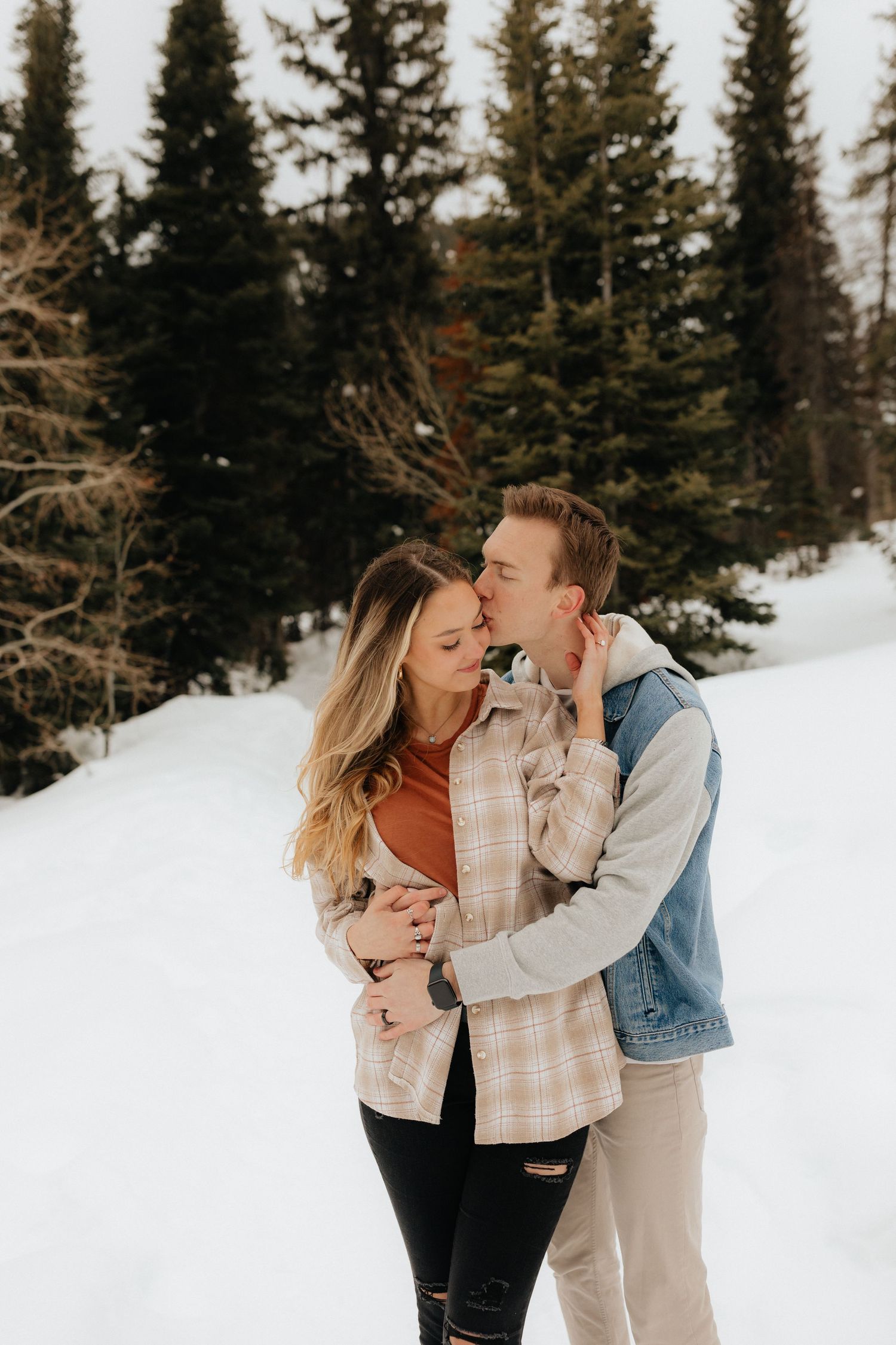 Man hugging woman from behind and kissing temple in snow filled mountains