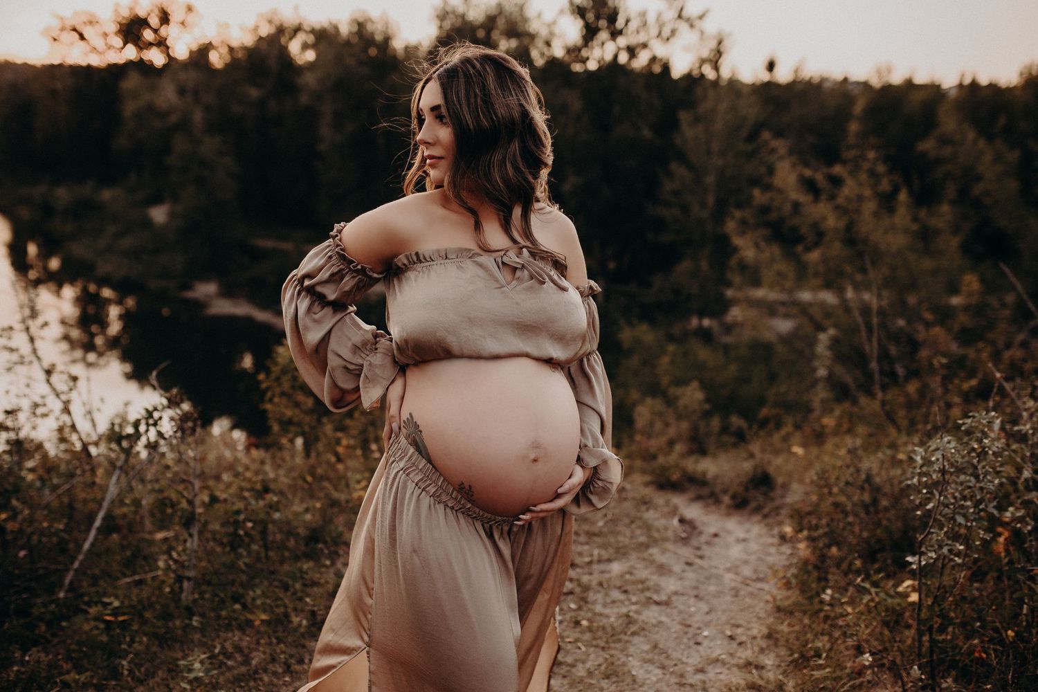 Silhouetted pregnant woman in flowing dress stands on nature path at sunset.