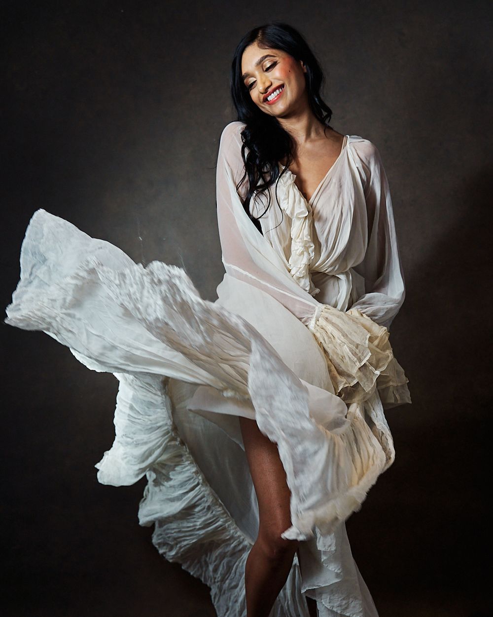 A woman in a flowing white dress smiles joyfully as fabric billows around her in a studio setting.