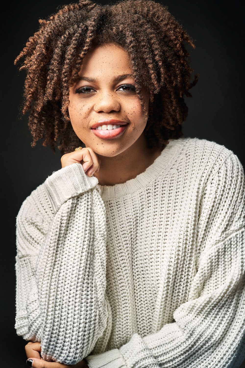 Young Black woman with natural curly hair and freckles smiles warmly in a white knit sweater.