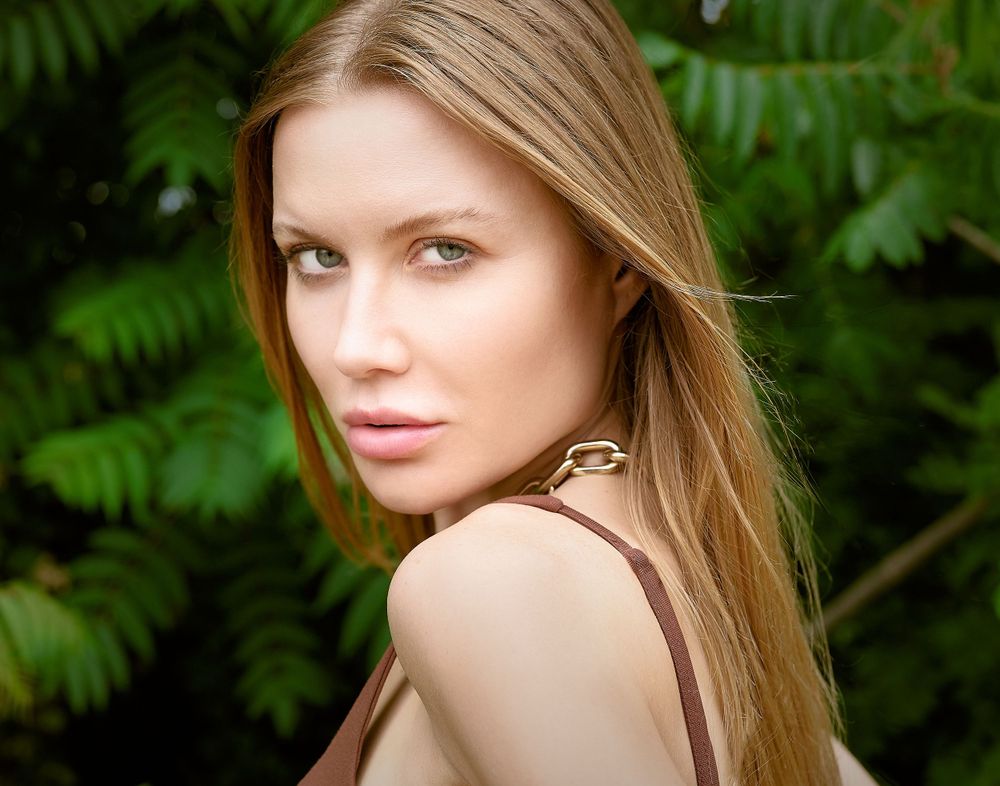 Blonde woman with long hair wearing brown top posing outdoors with green foliage background.