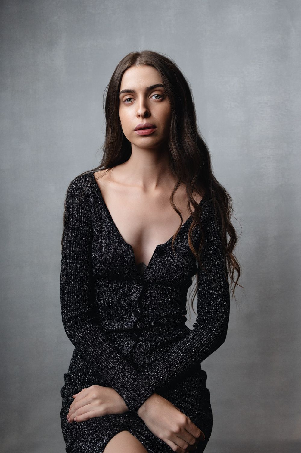 Brunette woman in a dark knit dress seated and posing against a gray textured studio background.