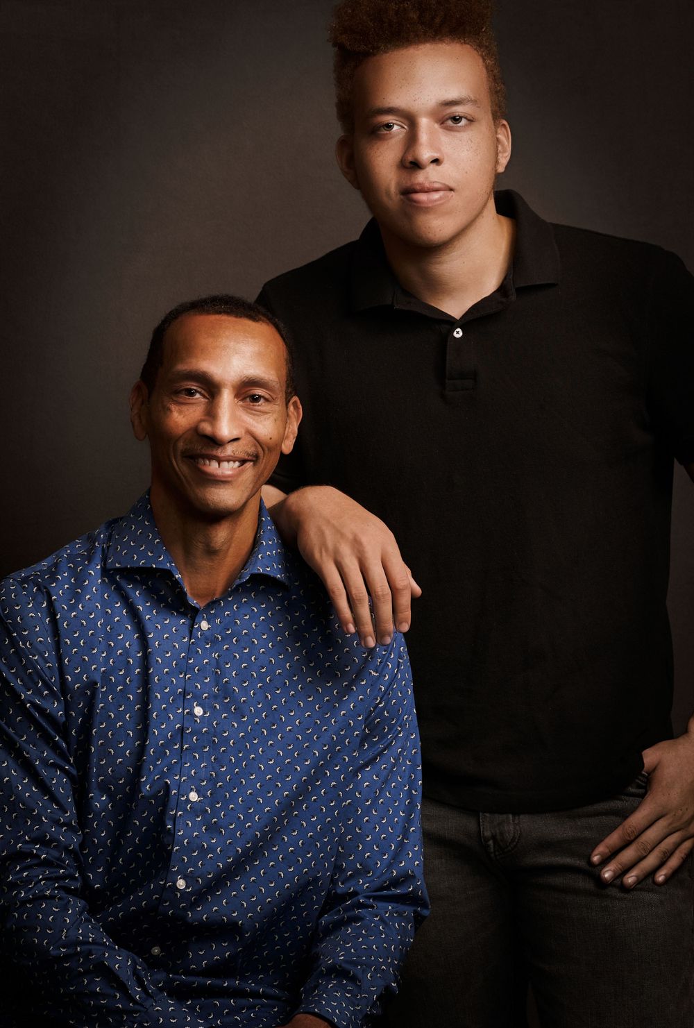 Two men, father and son, posing together for a portrait against a dark studio background.