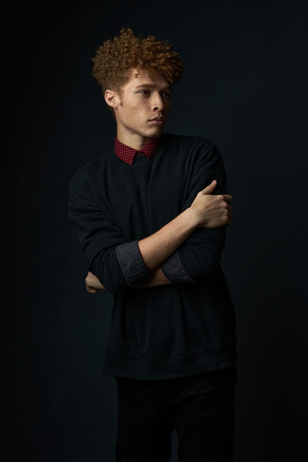 A young man with curly auburn hair stands with arms crossed wearing a black sweater with a red bow tie.