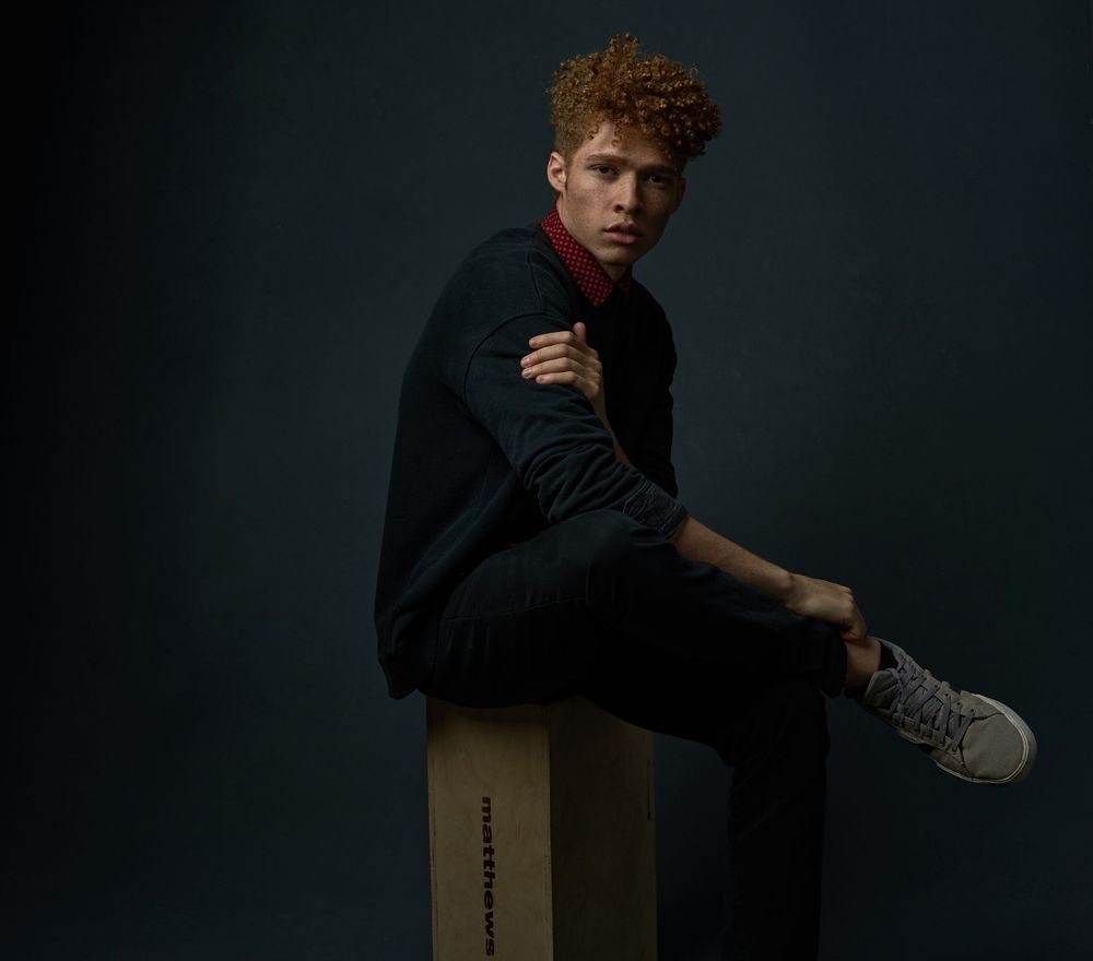 A young man with curly auburn hair sits on a box wearing a dark outfit against a moody teal background.