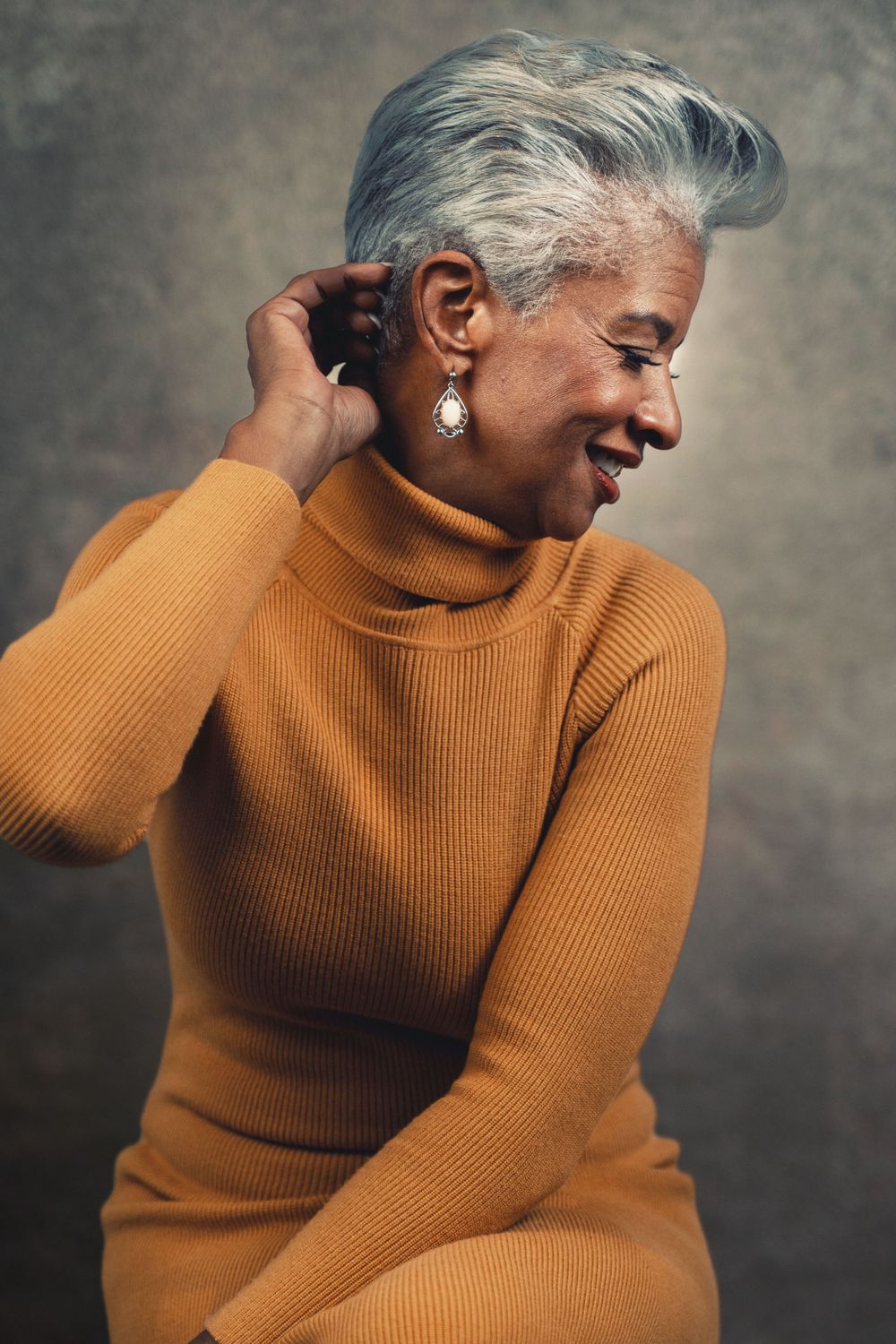 Stylish older woman with gray hair in an orange turtleneck sweater laughing in a studio portrait.