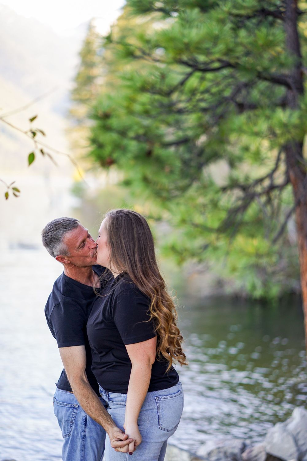 Romantic couple in matching black outfits embracing by a serene lake with trees in the background.