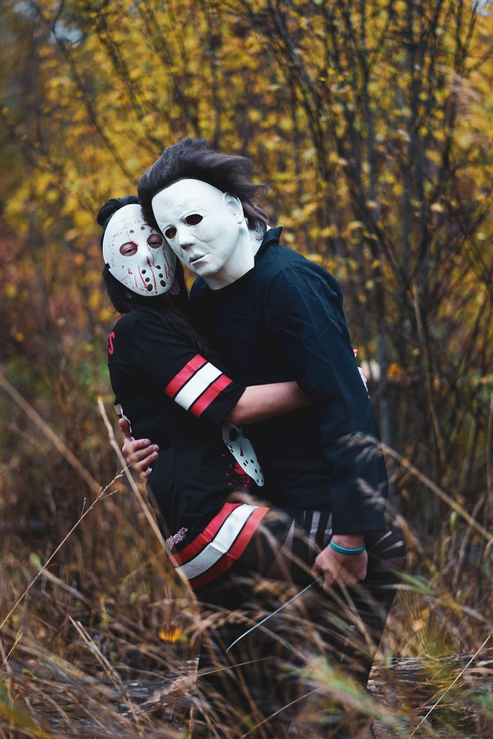 Two people in horror movie masks and dark clothing pose together among fall leaves.
