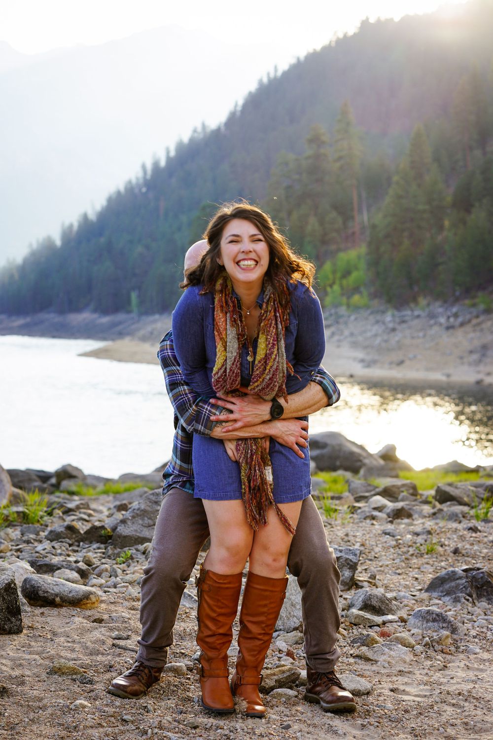 Couple shares playful moment on rocky shoreline with mountain vista behind.