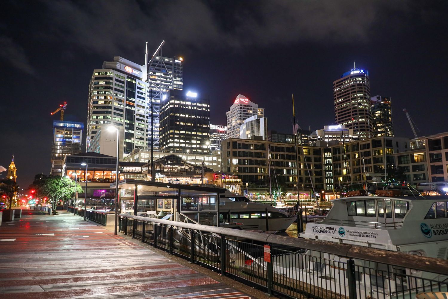 A bustling waterfront cityscape at night, featuring docked boats, illuminated high-rise buildings, and a boardwalk with railing in the foreg