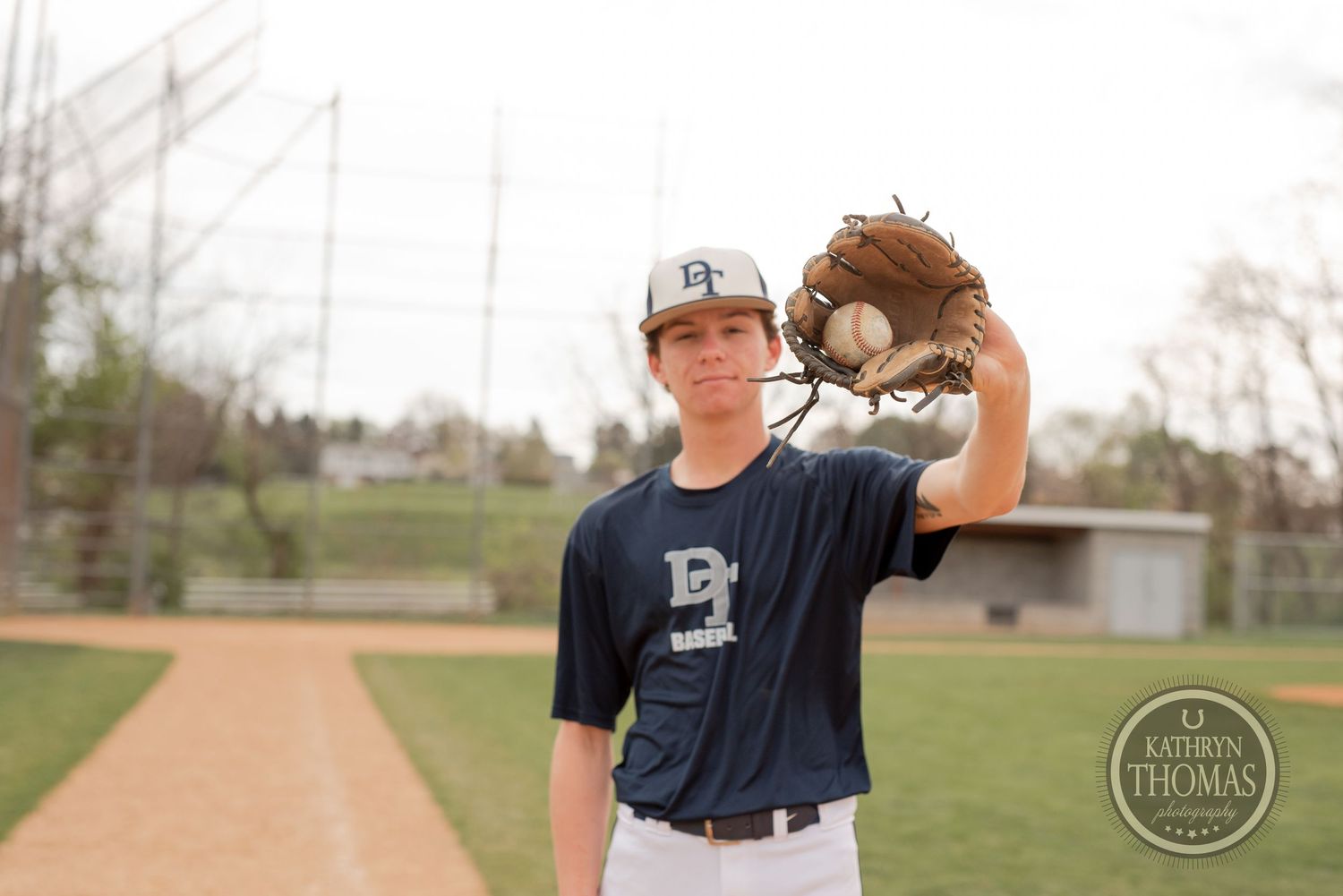 Baseball player in navy jersey holds up glove on baseball diamond with dirt path visible.