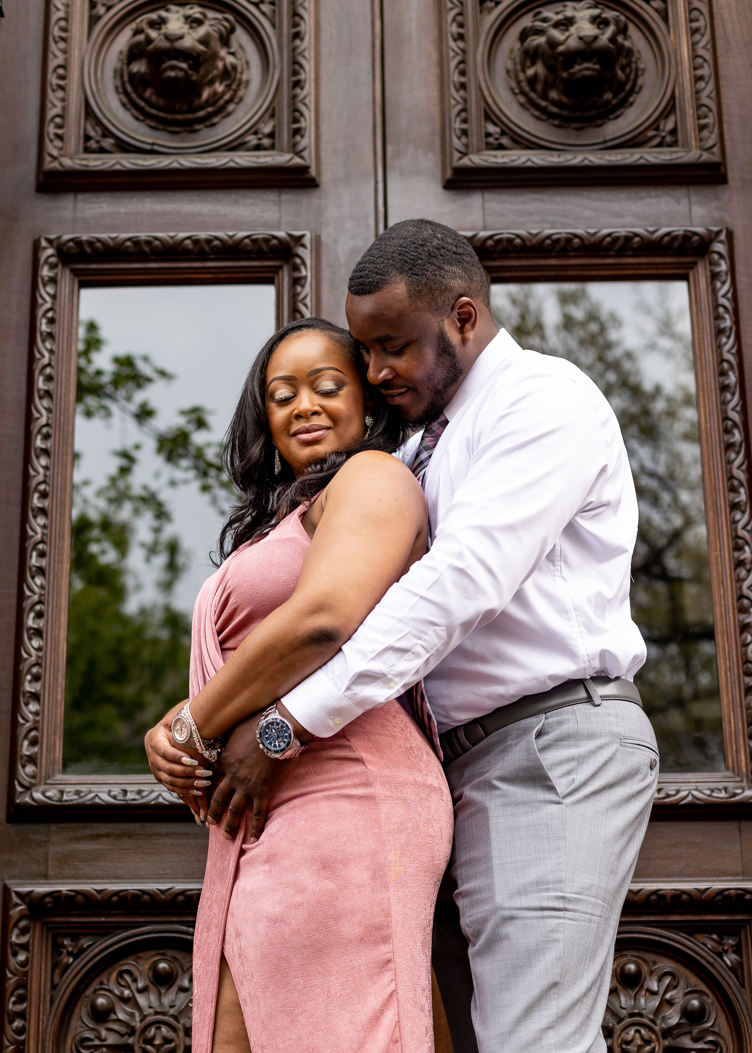 Romantic couple embracing before ornate wooden doors, woman in pink dress leaning into mans arms.