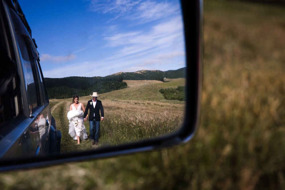 A side mirror reflection shows a couple in formal attire standing on a rural hillside next to their vehicle.