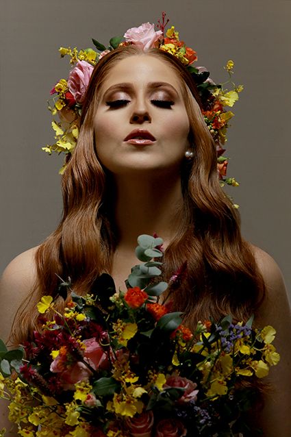 Artistic portrait featuring long wavy hair adorned with a floral crown and holding colorful wildflower bouquet against dark backdrop.