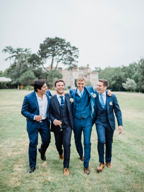 Un groupe de témoins de mariage en costumes bleu marine rit et marche ensemble à travers une pelouse verdoyante, avec un manoir en arrière-p