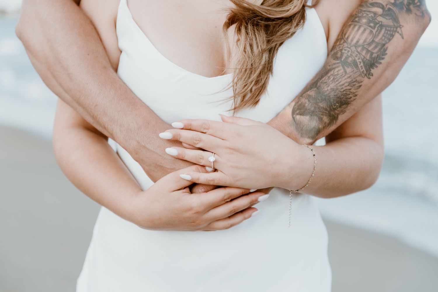 Close up of hands with engagement ring and tattoo embracing at the beach.