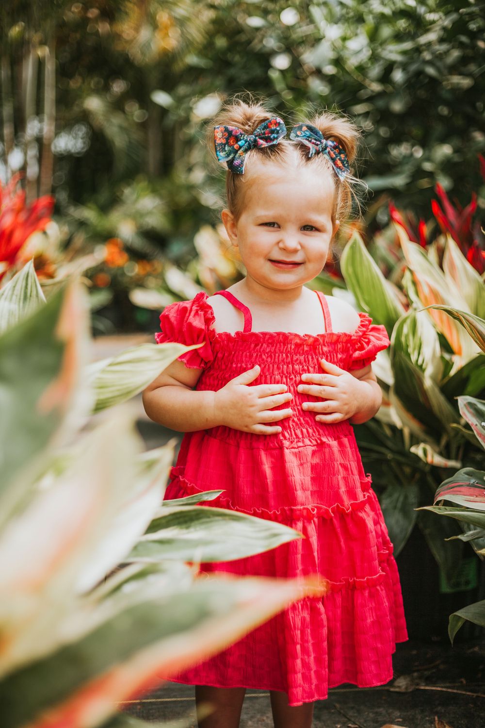 Child wearing a red dress smiles while standing in a lush tropical garden.