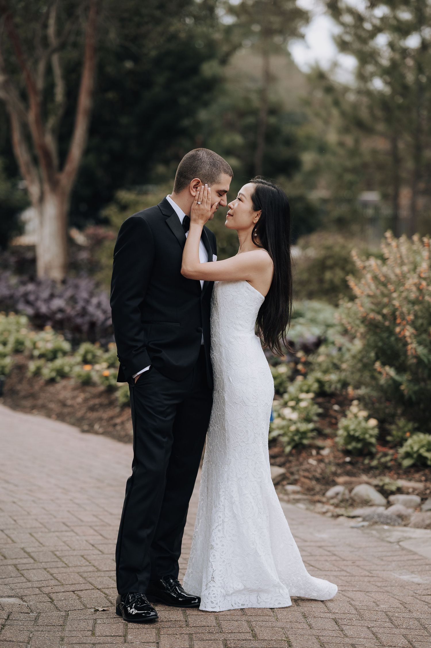 Elegant bride in fitted lace gown embraces groom on brick path surrounded by lush garden scenery.