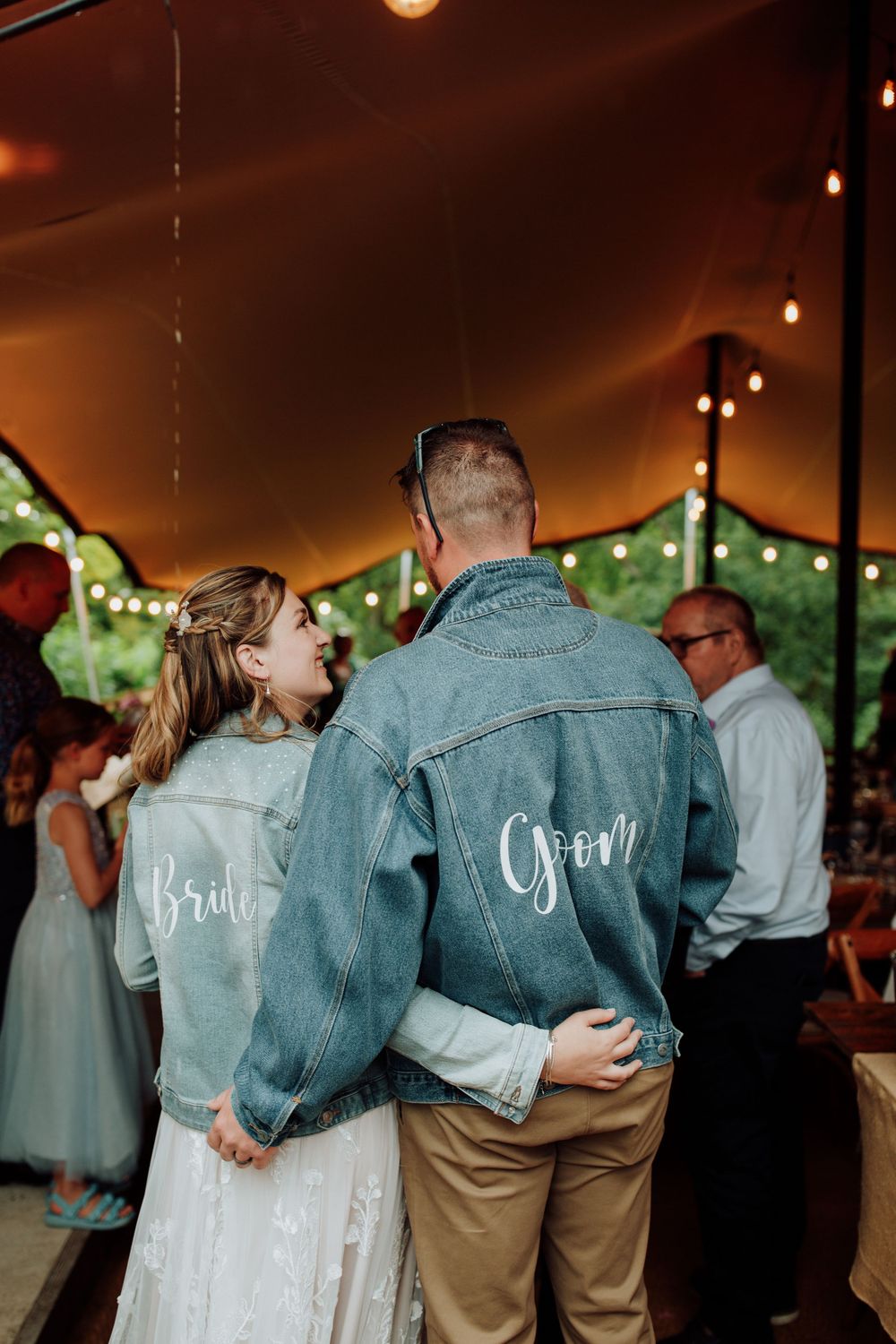 A couple wearing matching denim jackets with 'Bride' and 'Groom' text dance under string lights.