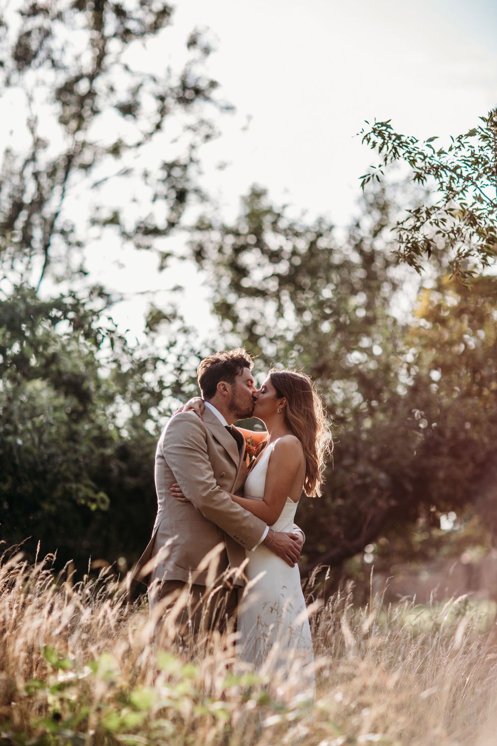 Romantic wedding couple embraces in tall grass during golden hour with dreamy backlit scenery.