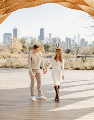 Couple holding hands walking on boardwalk with city skyline behind them.