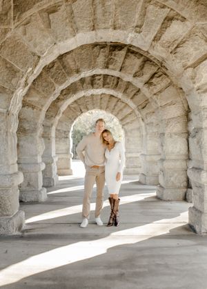 Stylish couple in neutral tones walking through stone archway tunnel with dramatic lighting.
