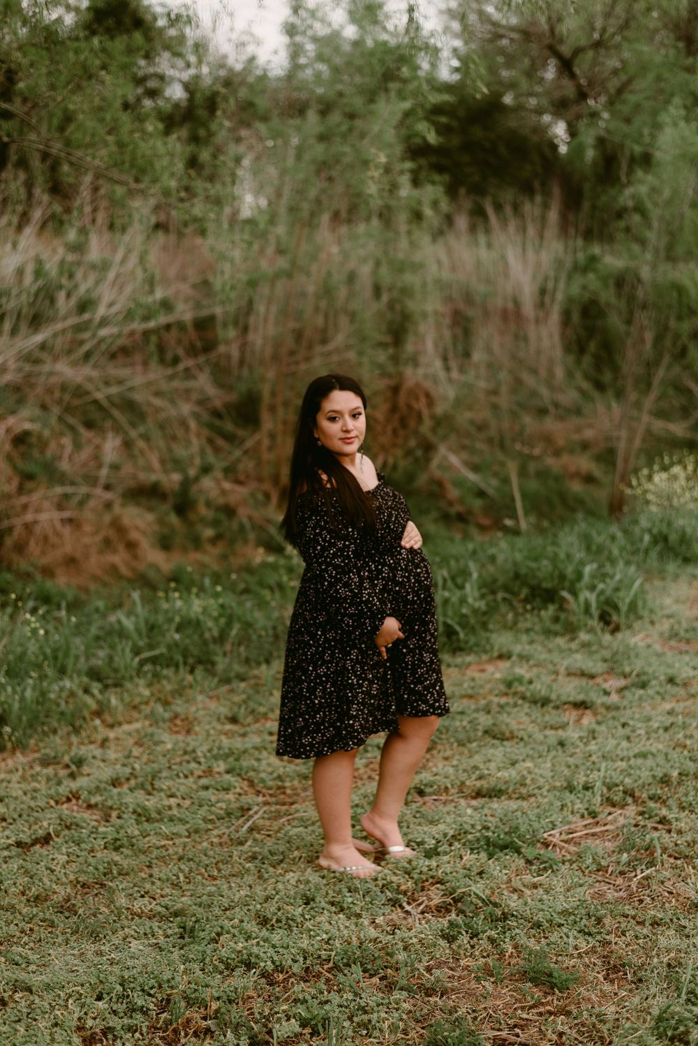 A woman in a black dotted dress stands barefoot in a wooded outdoor setting.