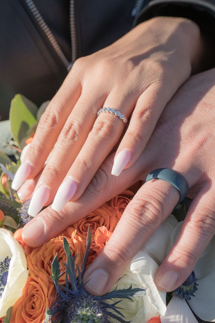 Close-up of newlyweds hands displaying wedding rings resting on a colorful bridal bouquet with orange and white roses.