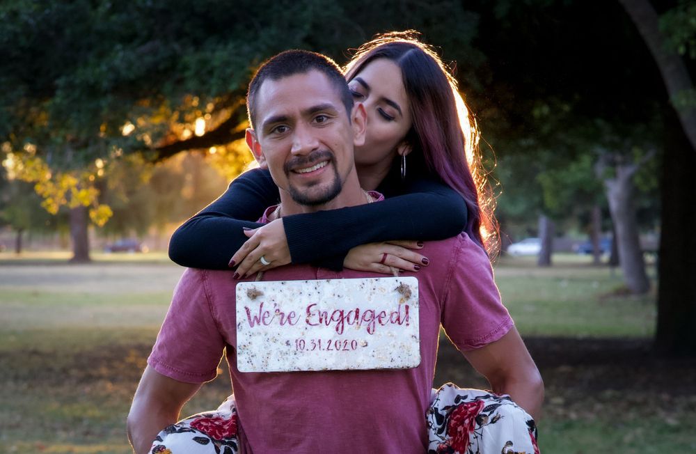Happy couple holding a We're Engaged sign outdoors in a park during golden hour engagement photo session.
