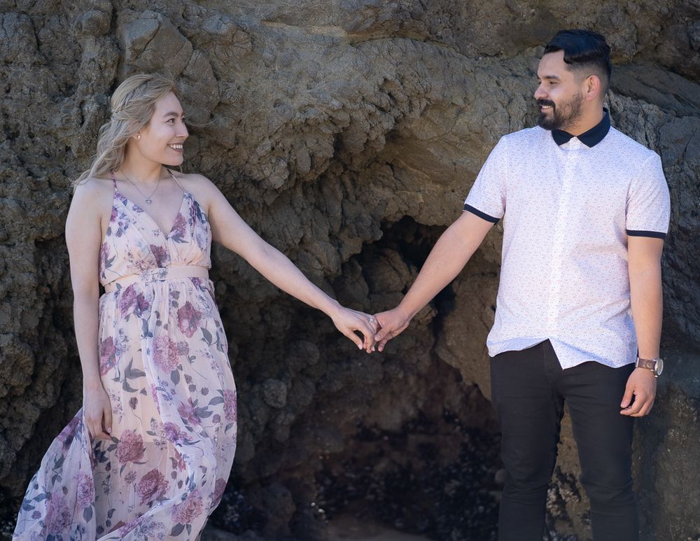 Couple holding hands in front of rocky cliffs, woman in floral maxi dress during beach engagement session.