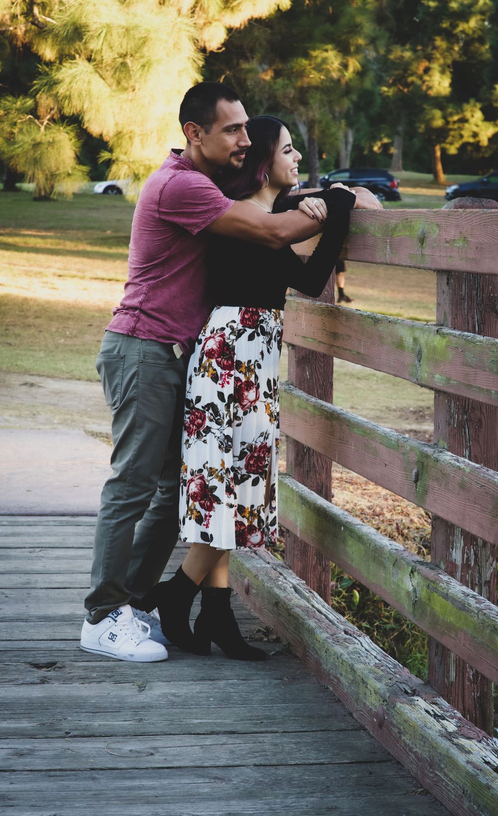 Engaged couple embracing on a wooden bridge in a park, woman wearing floral skirt during outdoor photo session.