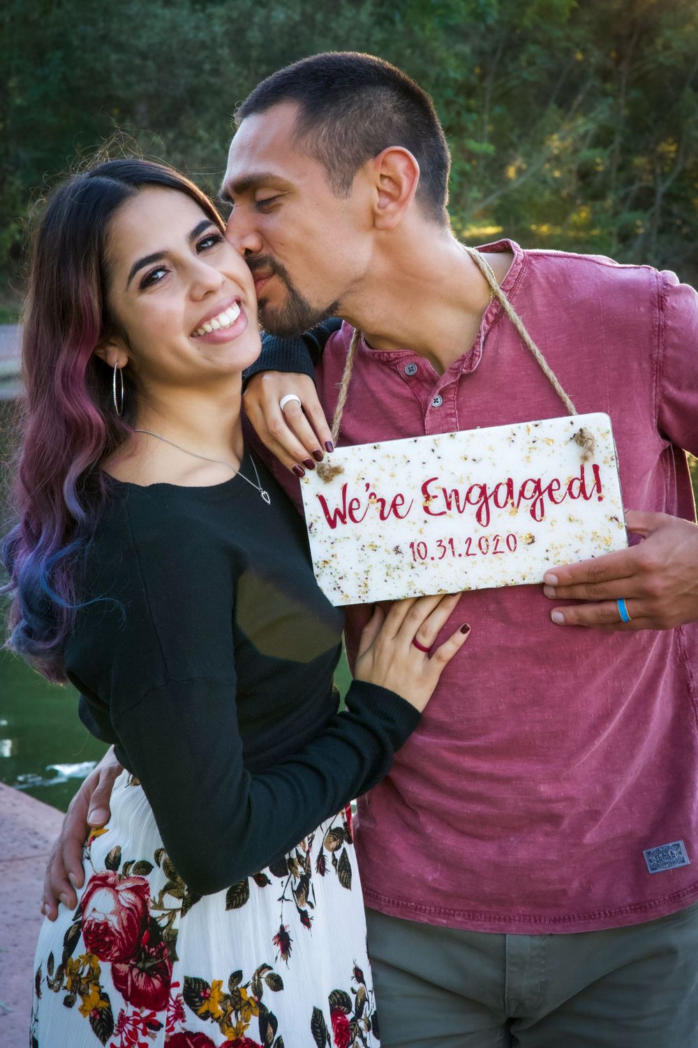 Smiling woman holds We're Engaged sign dated 10.31.2020 while partner kisses her cheek outdoors.