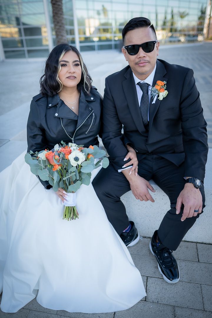 Bride in white gown with leather jacket and groom in black suit pose with bouquet outdoors.