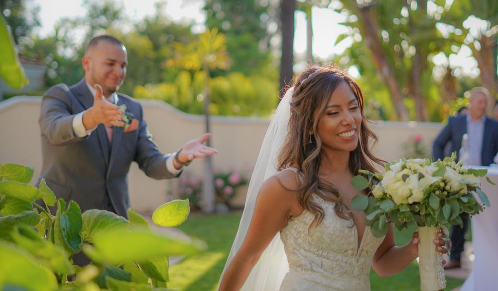 Radiant bride holding a white bouquet with groom gesturing behind her at an outdoor garden wedding.