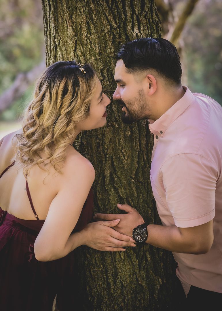 Romantic couple leaning in for a kiss against a large tree trunk in a serene outdoor park setting.