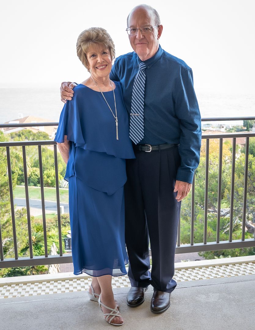 Elegant older couple dressed in coordinating blue outfits posing on a balcony with ocean views behind them.