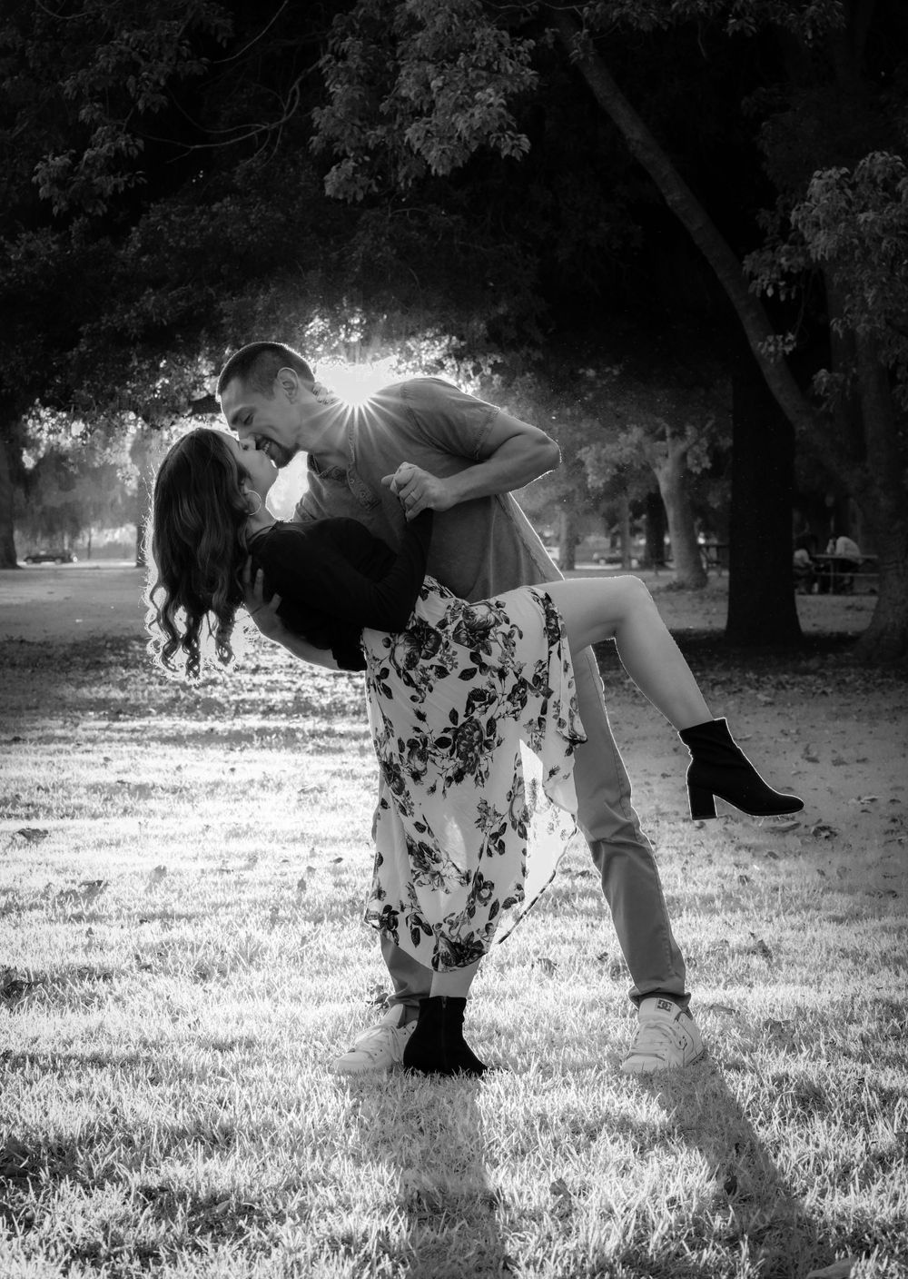 Romantic black and white photo of couple sharing a dip kiss in a sunlit park during engagement shoot.