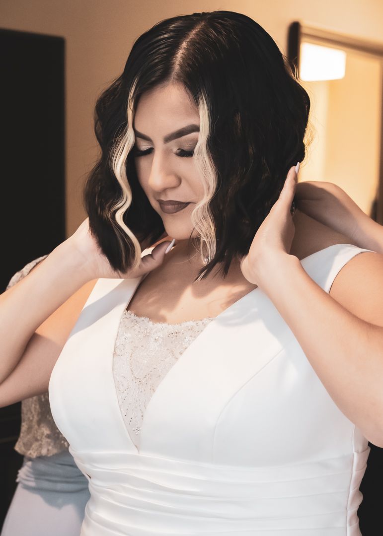 Bride in white wedding gown adjusts hair while getting ready for her wedding ceremony indoors.