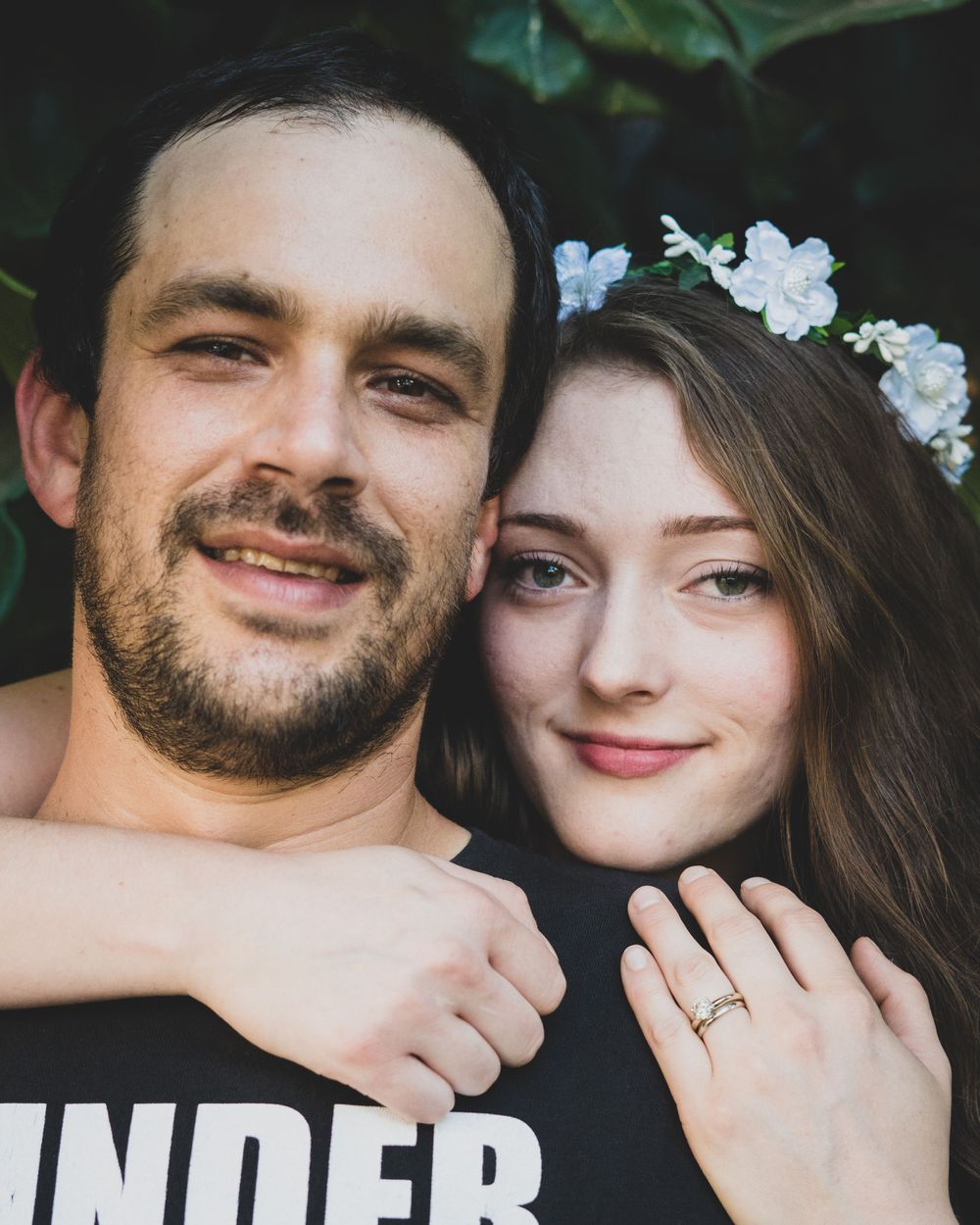 Smiling couple poses closely together outdoors, woman wearing floral crown and engagement ring in portrait.