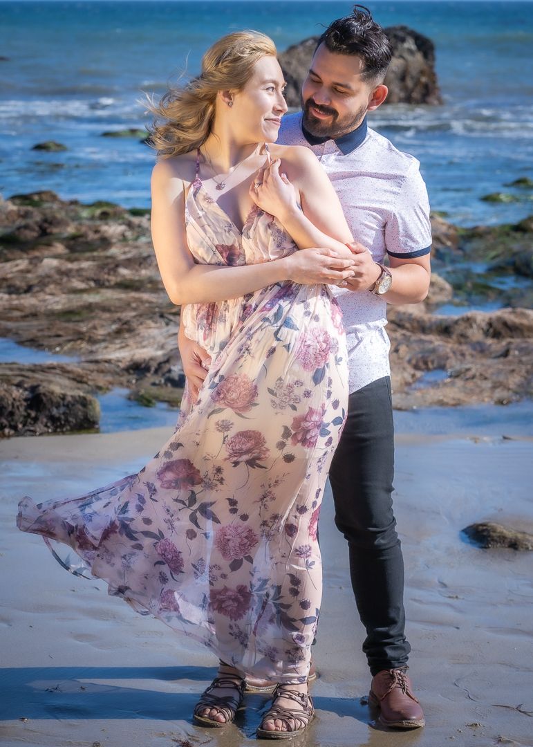Couple embracing on a rocky beach, woman in floral dress and man in white shirt, during engagement shoot.