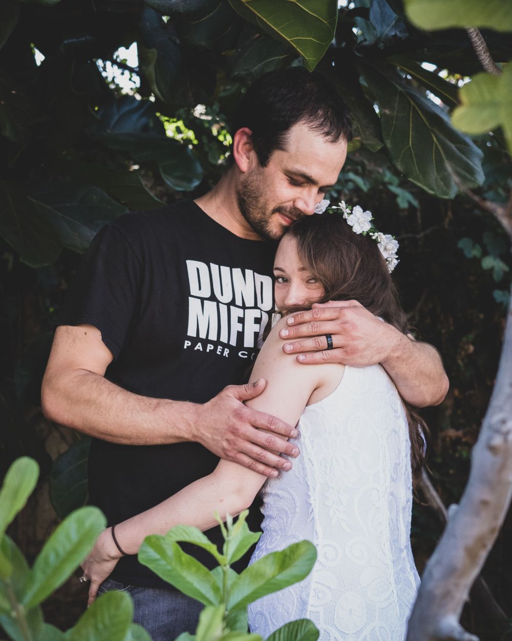 Man in Dunder Mifflin shirt tenderly embraces woman in white dress among tropical foliage in couples portrait.
