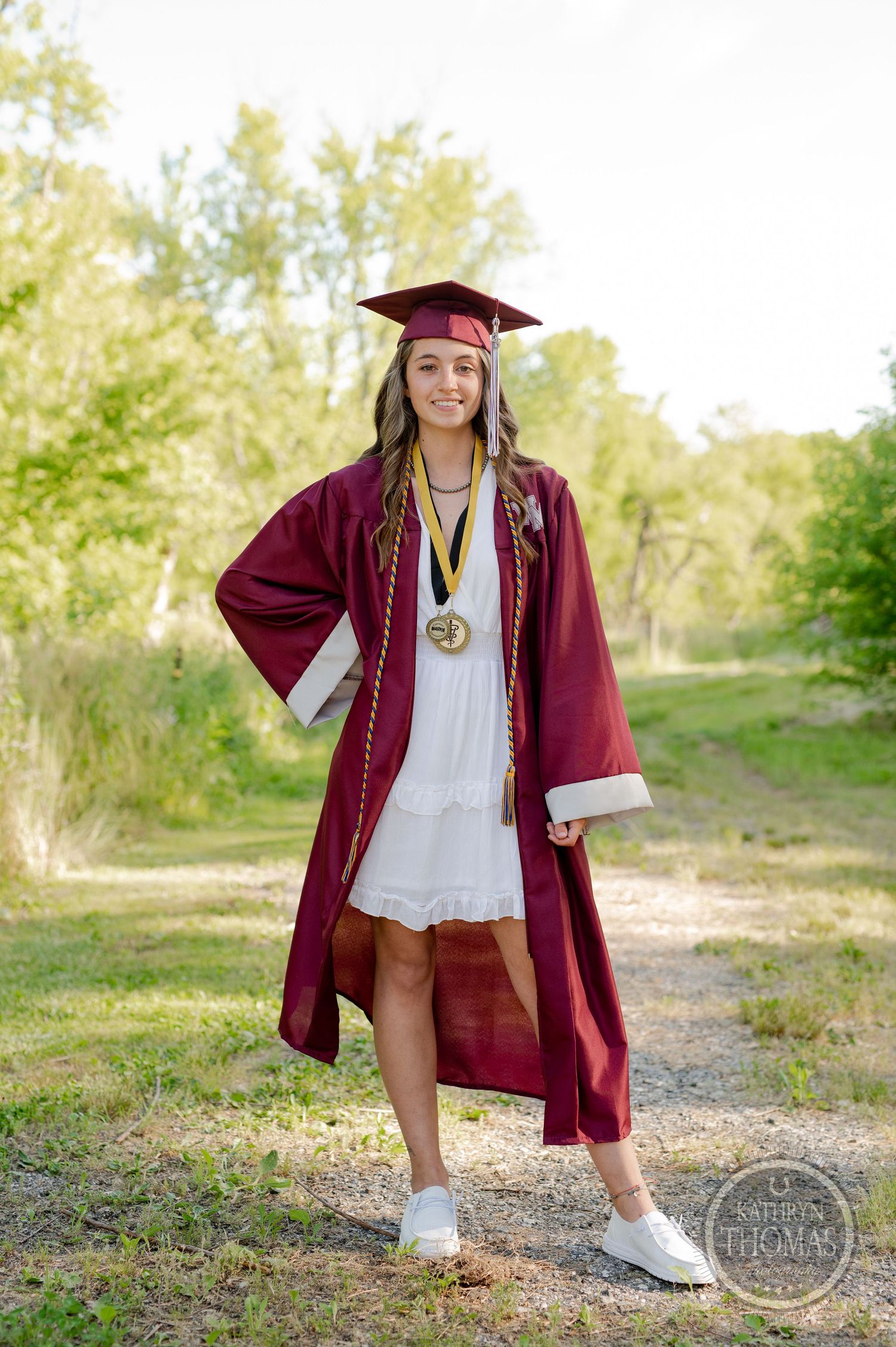 Graduate wearing maroon regalia and white dress poses on nature trail with graduation accessories.