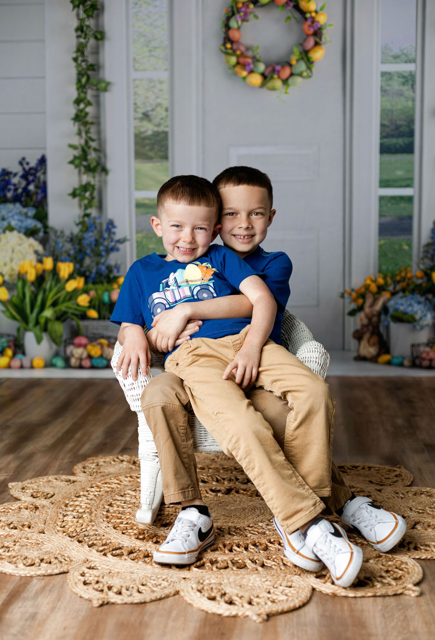 Two children sitting together on brown floor mat with floral wreaths in background.