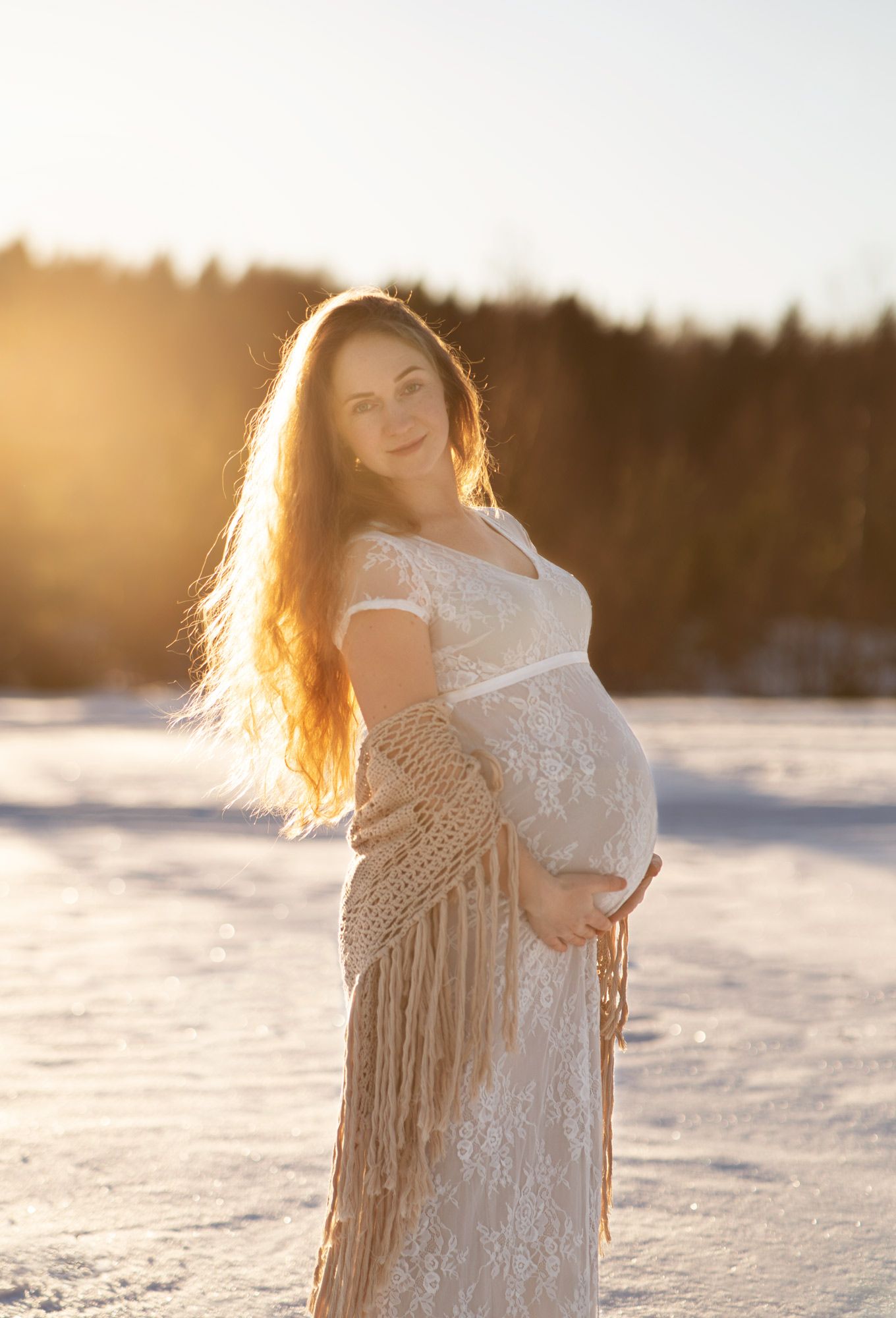 En långhårig gravid kvinna i en vit långklänning  med en beige sjal om armarna håller om sin mage. Solen lyser i hennes hår. Det är vinter.