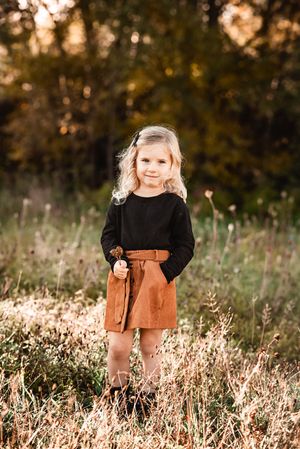 Young child in black top and brown skirt standing in golden sunlit field.